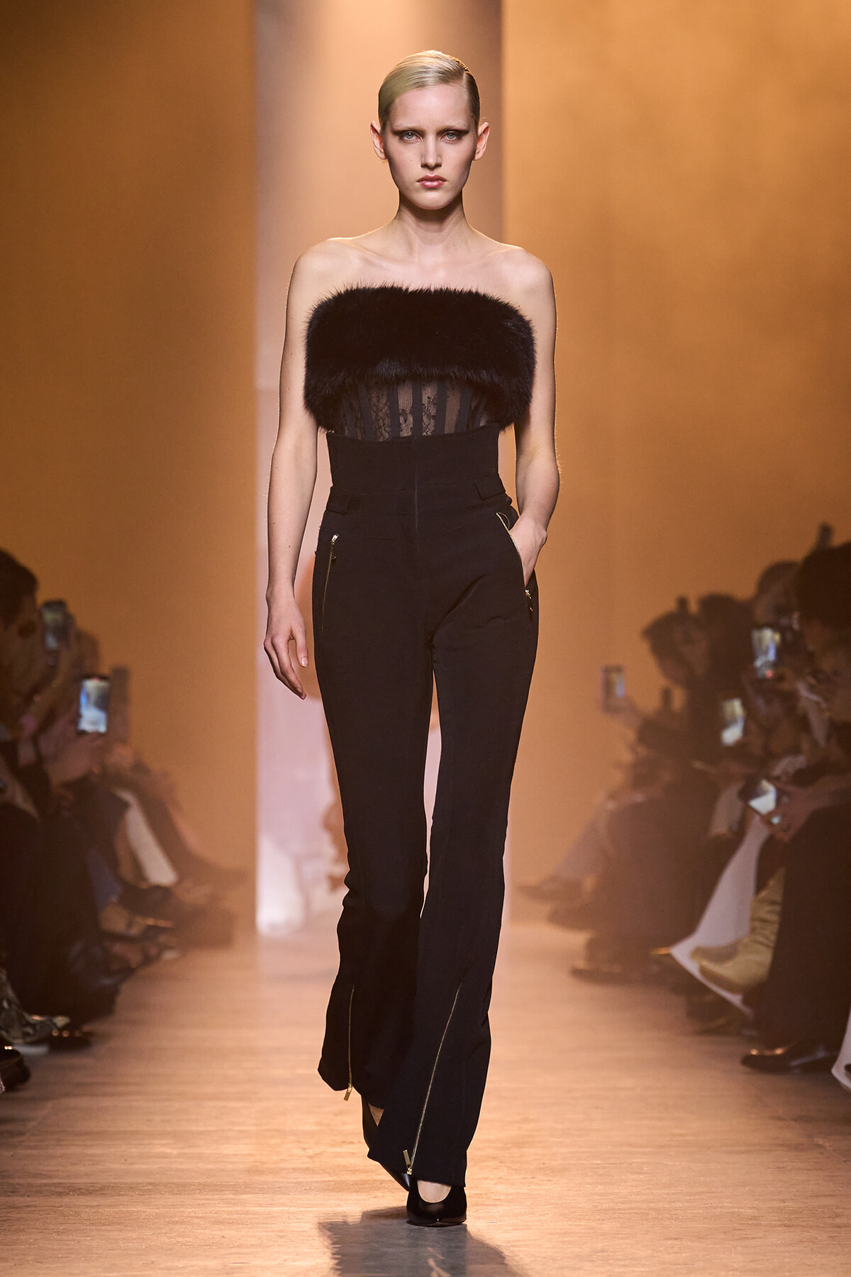Model walks the runway in a strapless black fur bustier and high-waisted black trousers with gold zippers, audience in the background