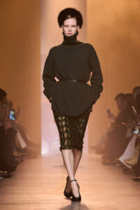 Model walks runway in oversized black turtleneck sweater belted at waist, paired with a sheer black lace pencil skirt and black heels under warm lighting.