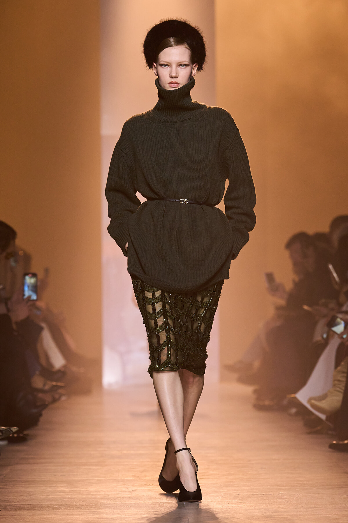 Model walks runway in oversized black turtleneck sweater belted at waist, paired with a sheer black lace pencil skirt and black heels under warm lighting.