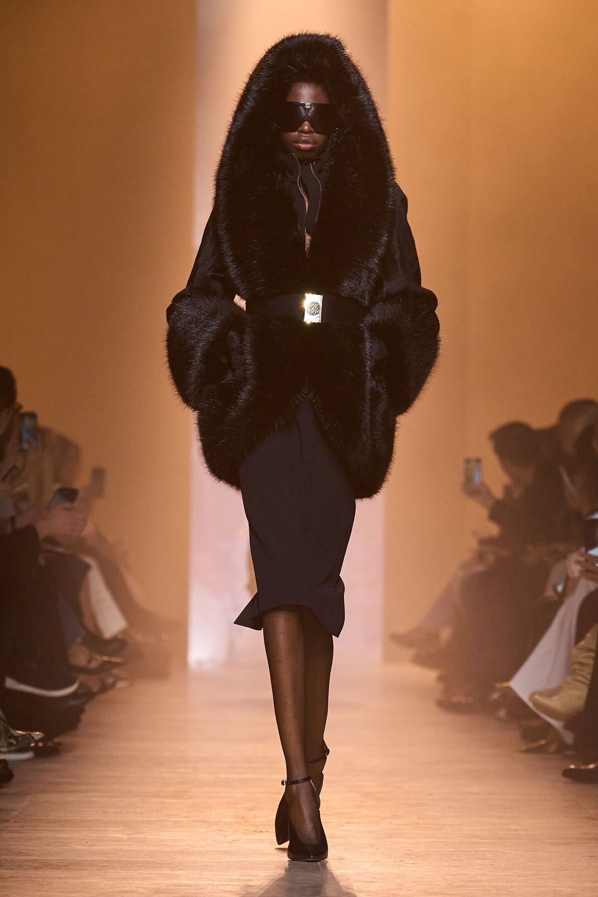 Model walks a runway in an oversized black fur coat with hood, black dress, and dark sunglasses, illuminated by warm lighting.