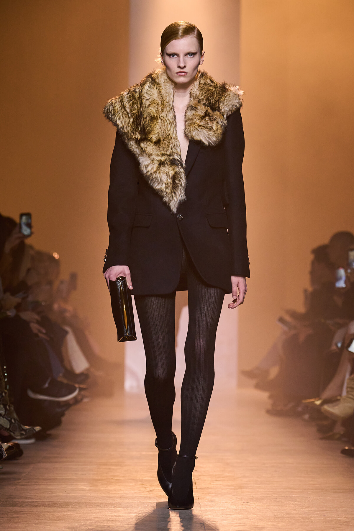 Model on a runway wearing a black blazer with a large fur collar, paired with striped tights, ankle boots, and a black clutch gesturing forward attitudes.