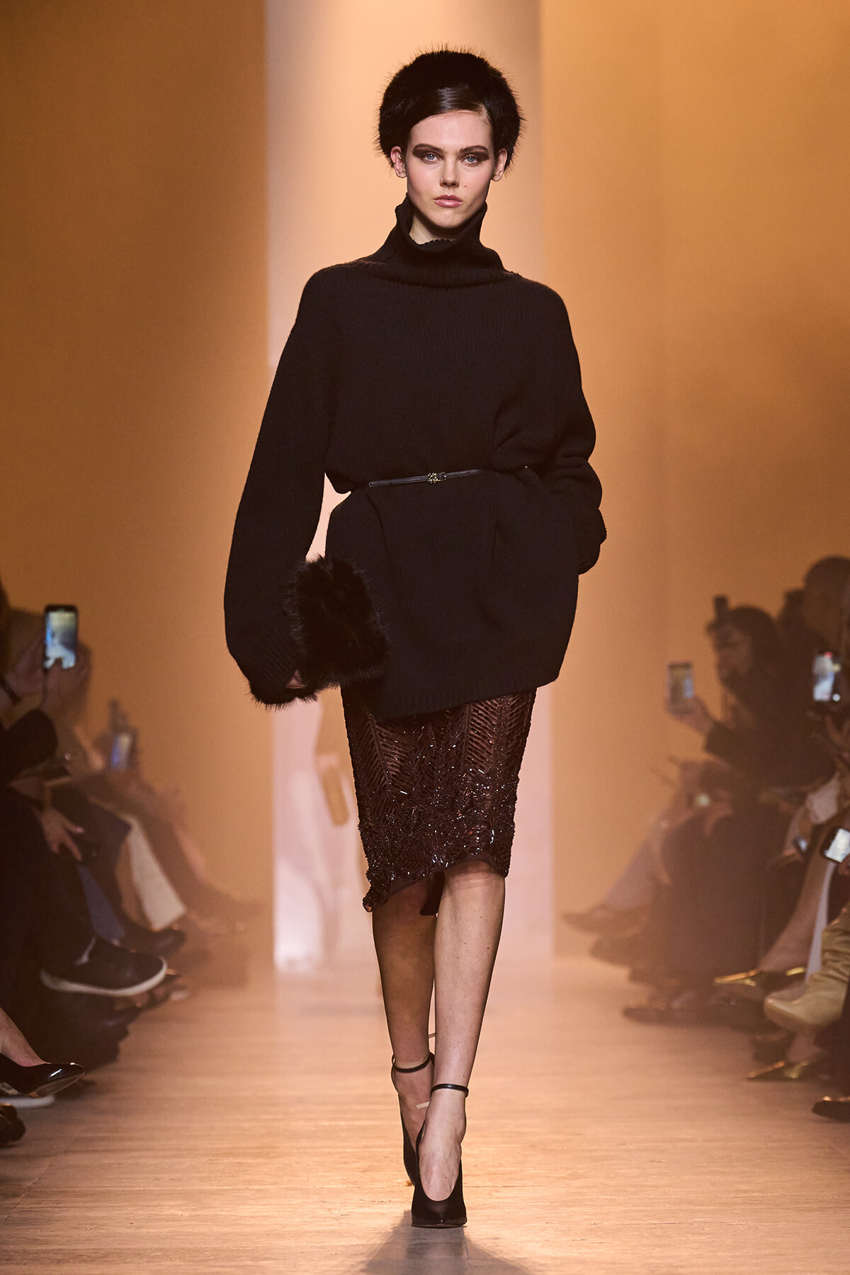 Model on a runway wearing an all-black ensemble: oversized turtleneck sweater with fur cuffs, a belted waist, and a sequined midi skirt with black heels.