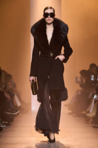 Model walks a fashion runway in a black fur-curred coat with a voluminous collar and sheer black pants, wearing oversized sunglasses and a small clutch.