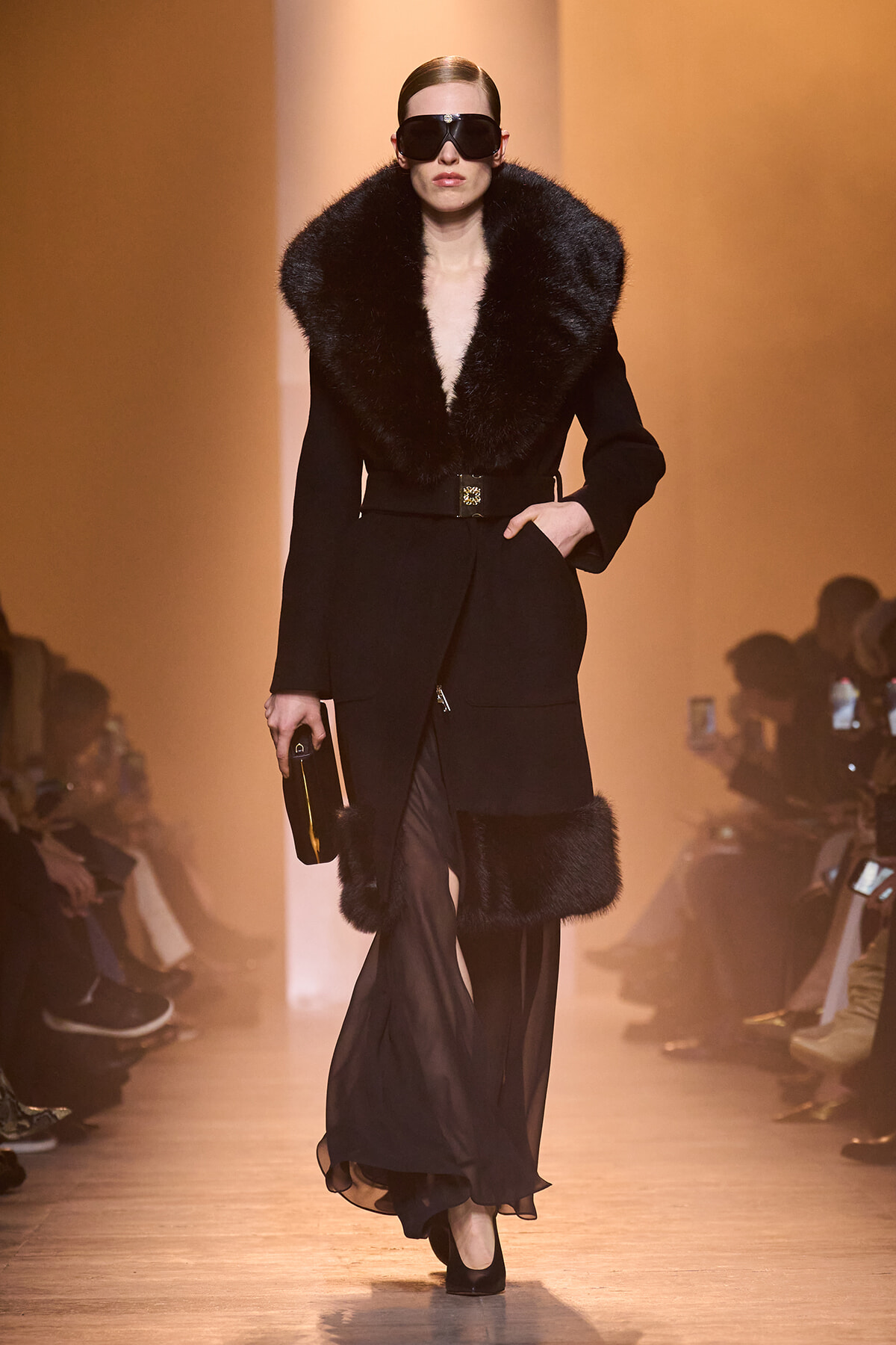 Model walks a fashion runway in a black fur-curred coat with a voluminous collar and sheer black pants, wearing oversized sunglasses and a small clutch.
