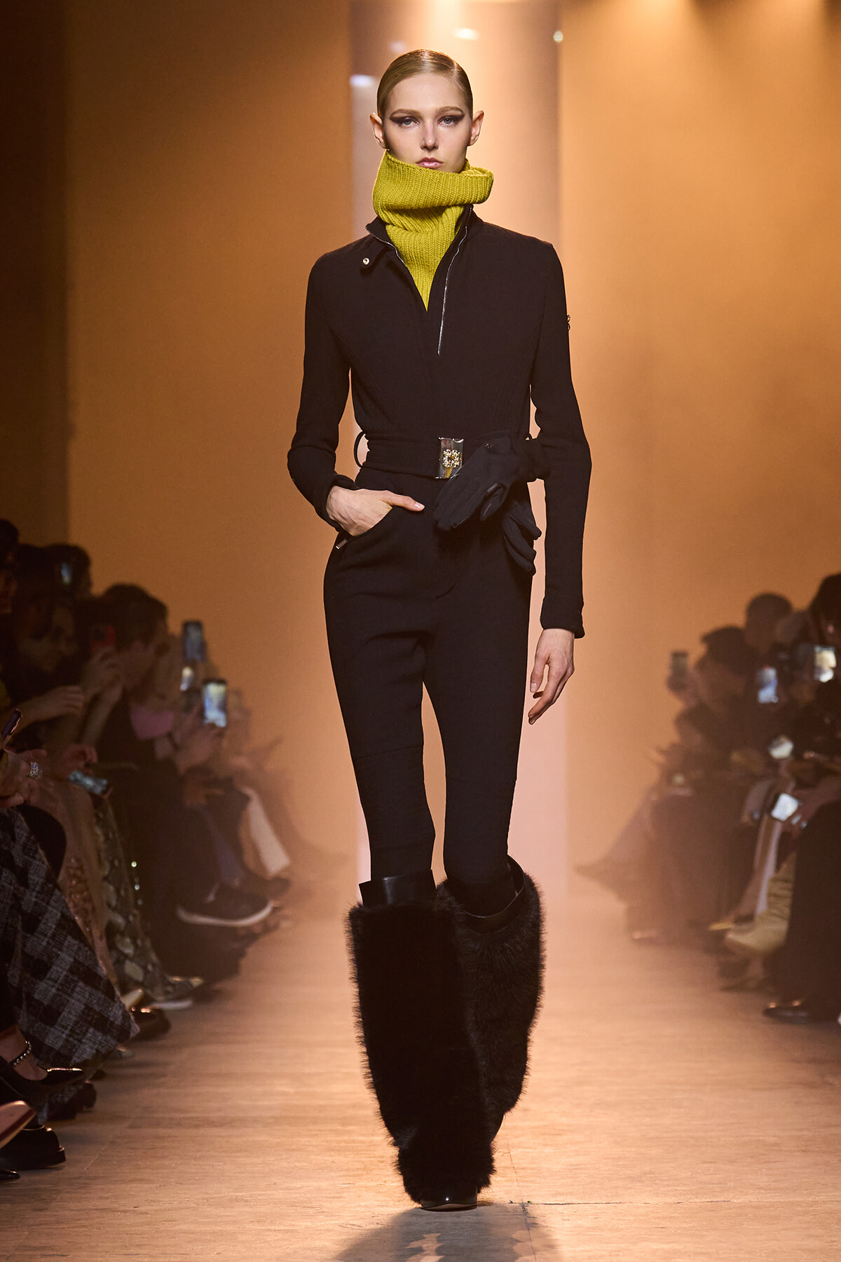 Model walks the runway in a black jumpsuit with a bright yellow scarf, belted waist, and oversized black fur boots.