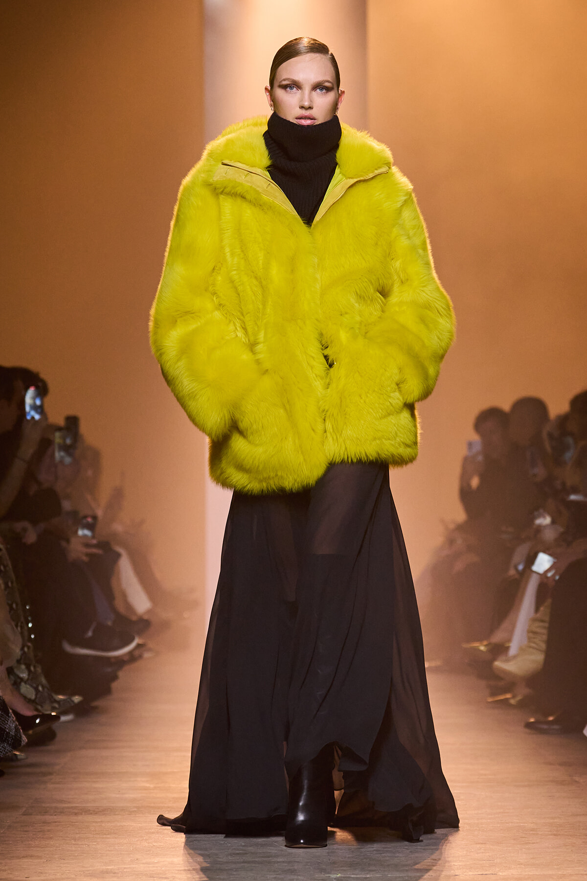 Model on a fashion runway wearing a bright yellow fur coat over a black turtleneck and long black skirt with audience in the background.