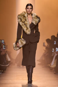 Model walks the runway in a black tailored suit with a large tan fur collar and cuffs, holding a matching fur accessory.