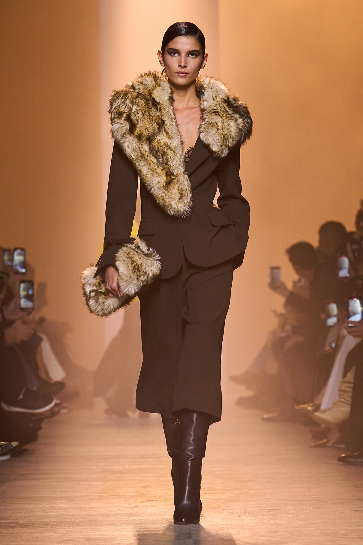 Model walks the runway in a black tailored suit with a large tan fur collar and cuffs, holding a matching fur accessory.