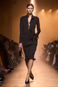 Model in a black tailored zip-front dress with a belt and gloves walking a fashion runway under warm spotlight.