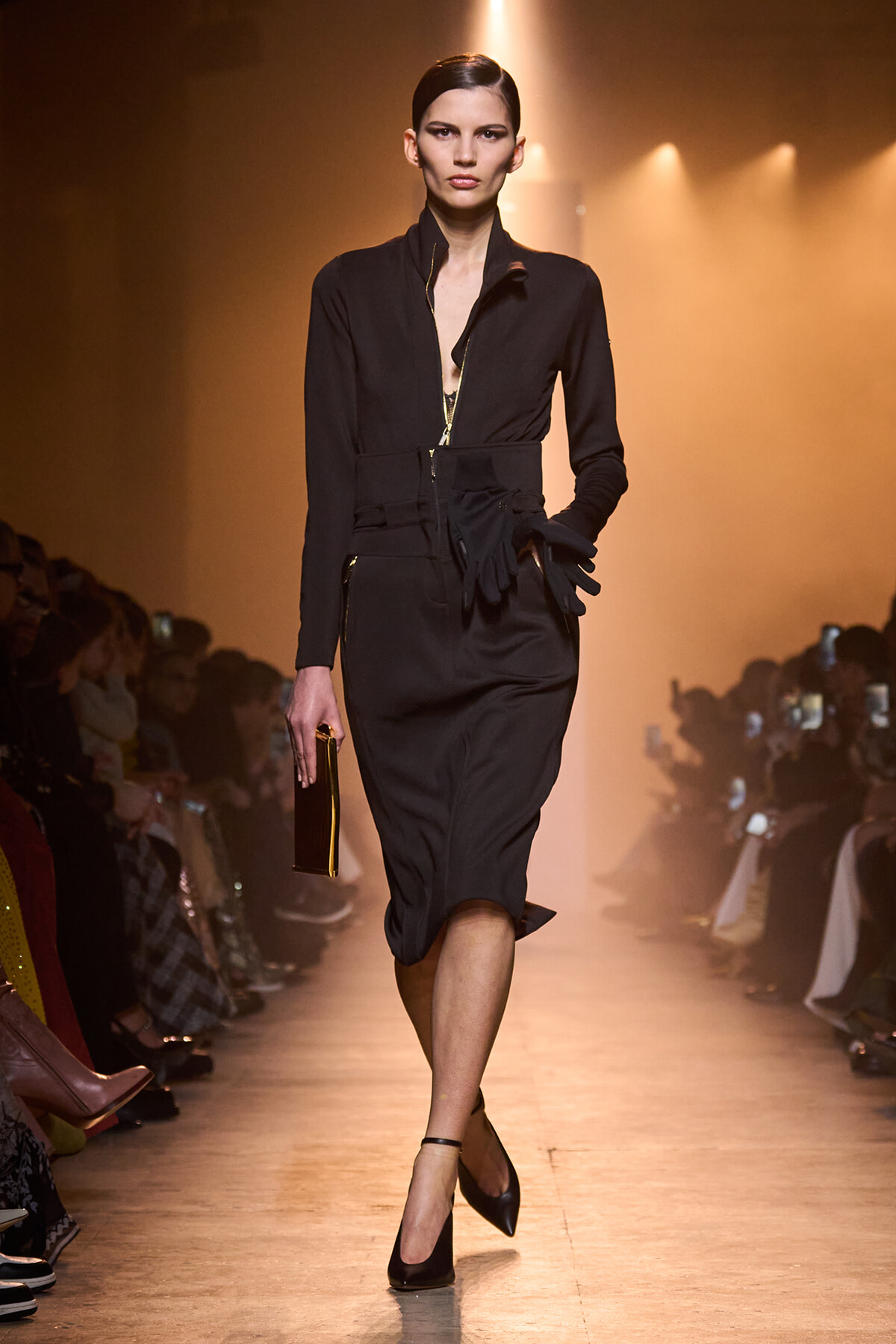 Model in a black tailored zip-front dress with a belt and gloves walking a fashion runway under warm spotlight.