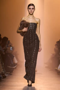 Model walks a black beaded strapless gown with vertical sequins, holding a brown fur stole on the runway.