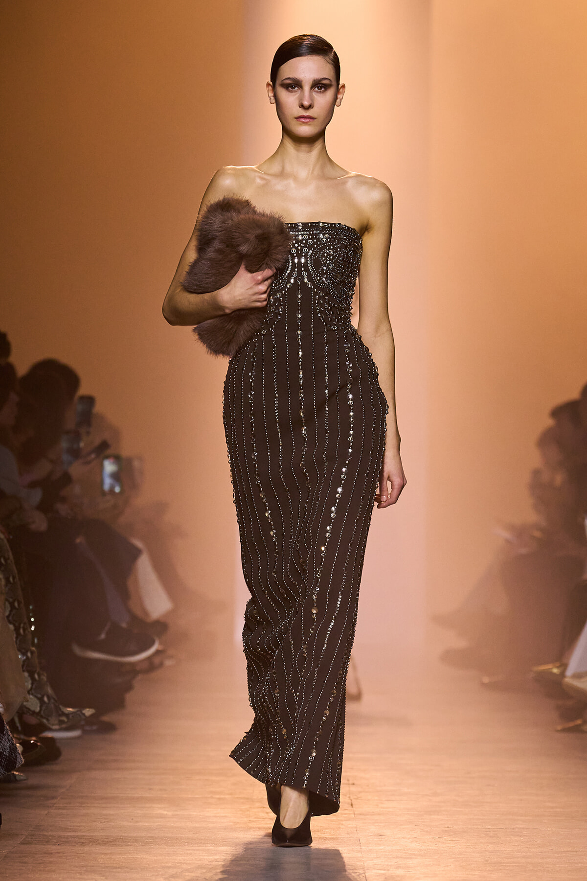 Model walks a black beaded strapless gown with vertical sequins, holding a brown fur stole on the runway.