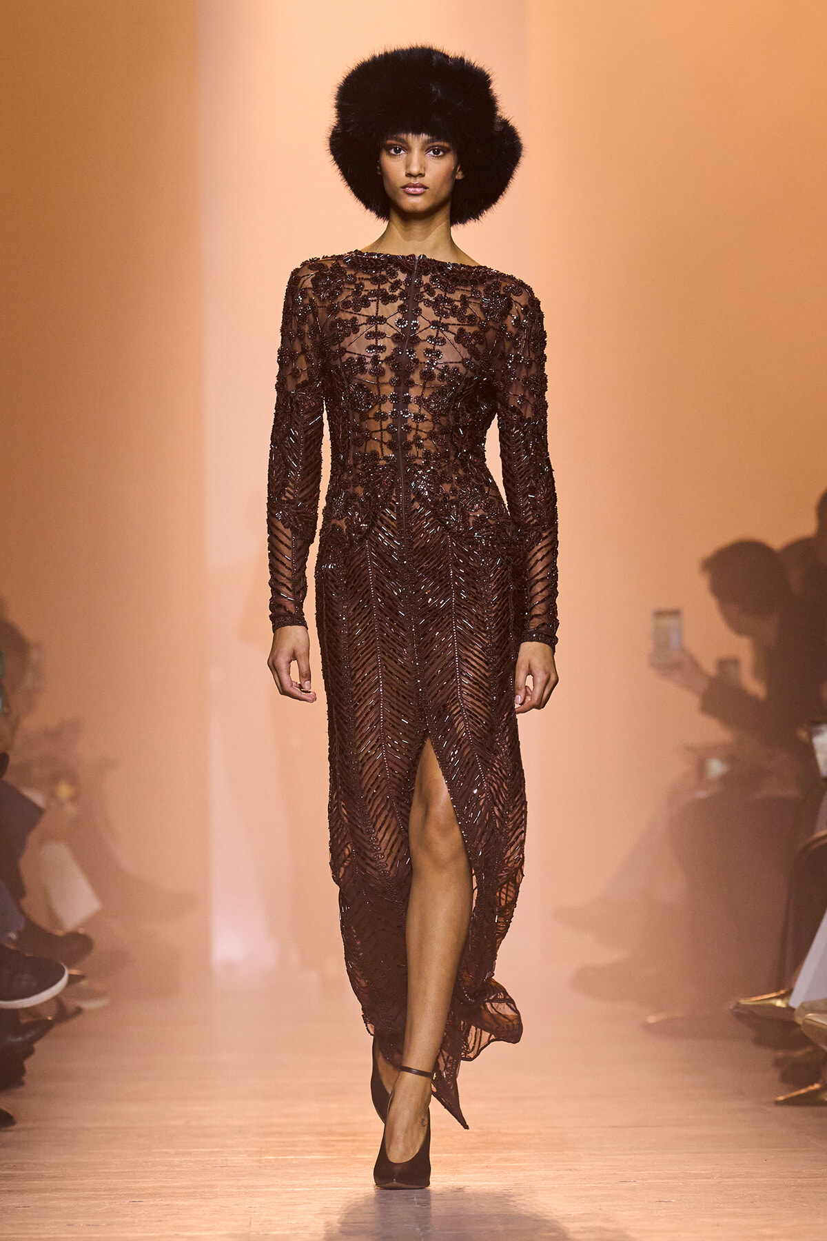 Model walking a runway in a sheer burgundy beaded gown with long sleeves, a high neckline, and a front slit, paired with a black fur hat and black heels against an orange backdrop.