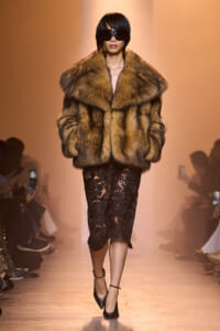 Model on a runway wearing a large brown fur coat over a black lace midi dress and black heels with oversized sunglasses.