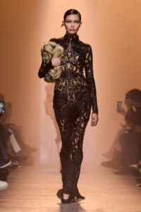 Model walking a runway in a black, intricate lace gown with sheer panels, holding a fur stole over her left shoulder.