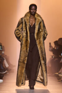 Model walks the runway in a long brown fur coat over a dark brown jumpsuit, hands in coat pockets against a warm orange backdrop.