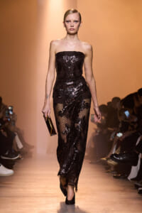 Model on runway wearing a strapless black sequin lace gown, holding a gold clutch, walking toward camera.