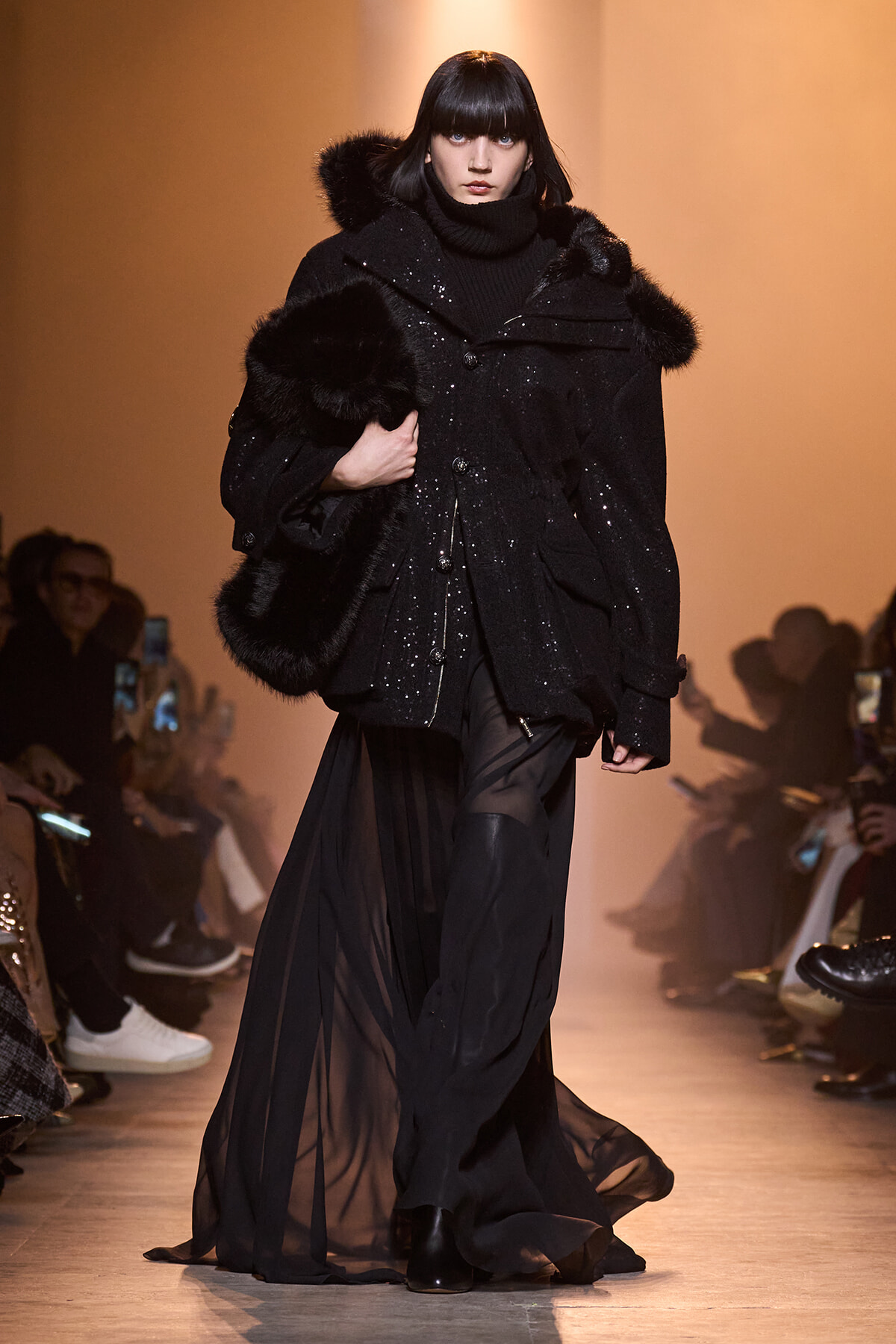 Model walks the runway in an oversized black coat with fur trim and a sheer, floor-length skirt, wearing a black turtleneck.
