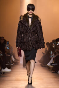 Model walks a runway in a dark sequin coat with fur collar, oversized sunglasses, black knee-length skirt, black heels, and a burgundy clutch.