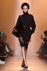 Model on a runway wearing a black knit turtleneck sweater dress with a slim belt, lace stockings, and black ankle-strap heels, carrying a large black fur bag.