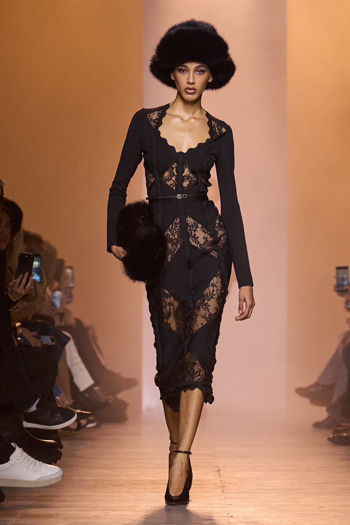Model walking a fashion show in a black lace dress with a matching fur hat and clutch against an warm orange backdrop.