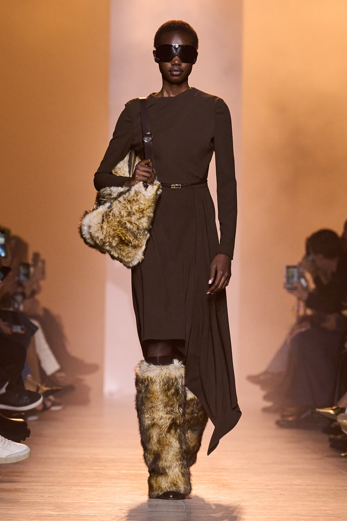 Model walking a runway in a dark, long-sleeve dress with a belt, paired with tall fur boots and a large fur bag slung over the shoulder, wearing oversized sunglasses.