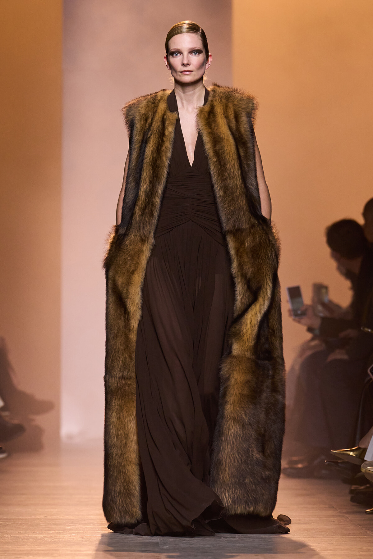 Model walks a runway in a full-length brown fur coat over a dark brown, ruched gown; warm orange backstage lighting surrounds the scene.