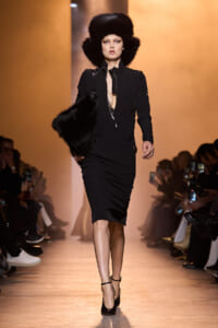 Model on a fashion show runway wearing a black zip-front dress with a dramatic oversized black fur hat and matching fur stole, audience in the background.