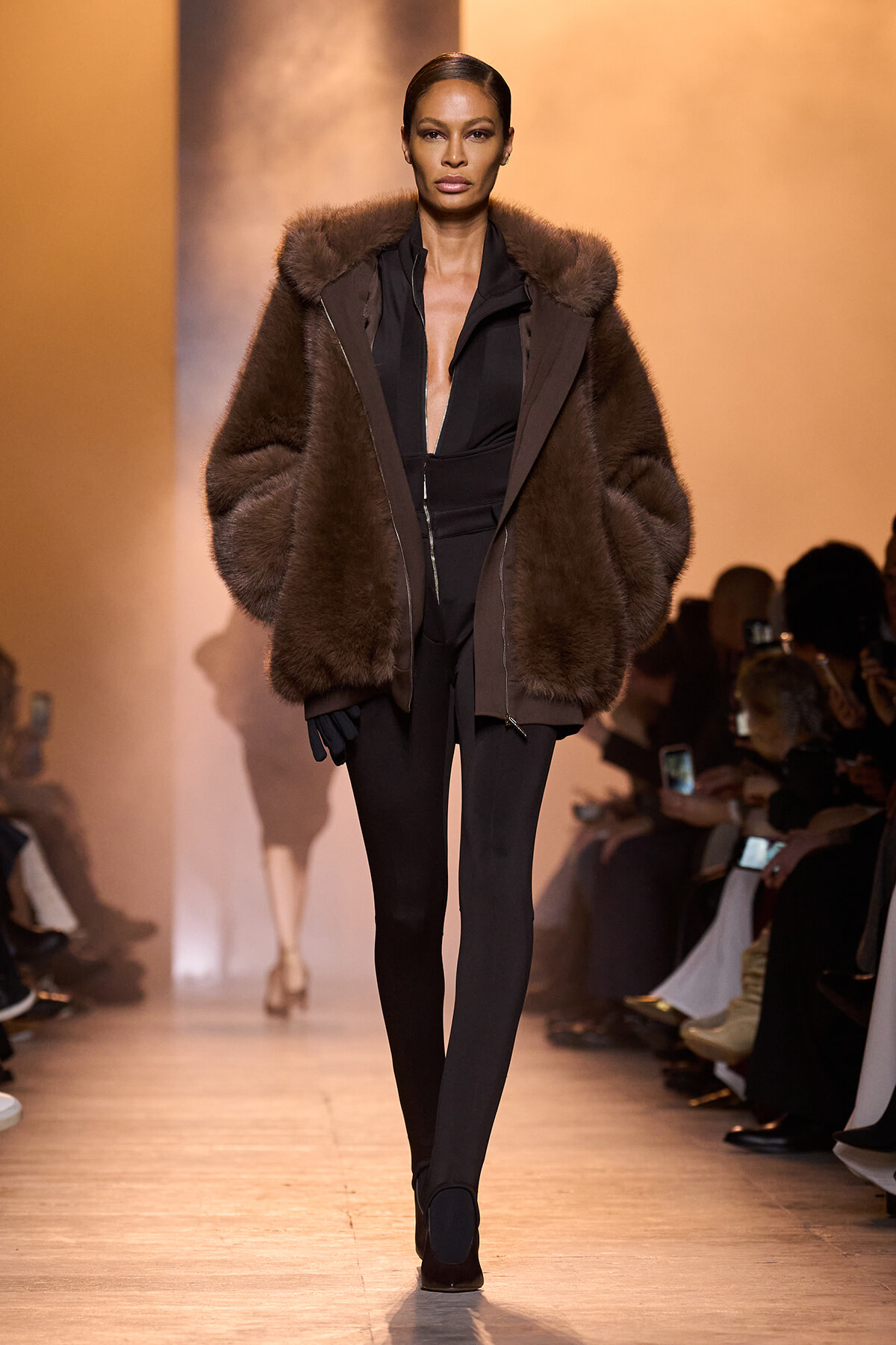 Model on runway wearing a large brown fur coat over a black zip-front jumpsuit, with black gloves and heels.