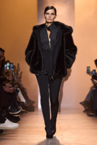 Model walking the runway in an all-black outfit with a large black fur coat, audience watching and filming nearby.