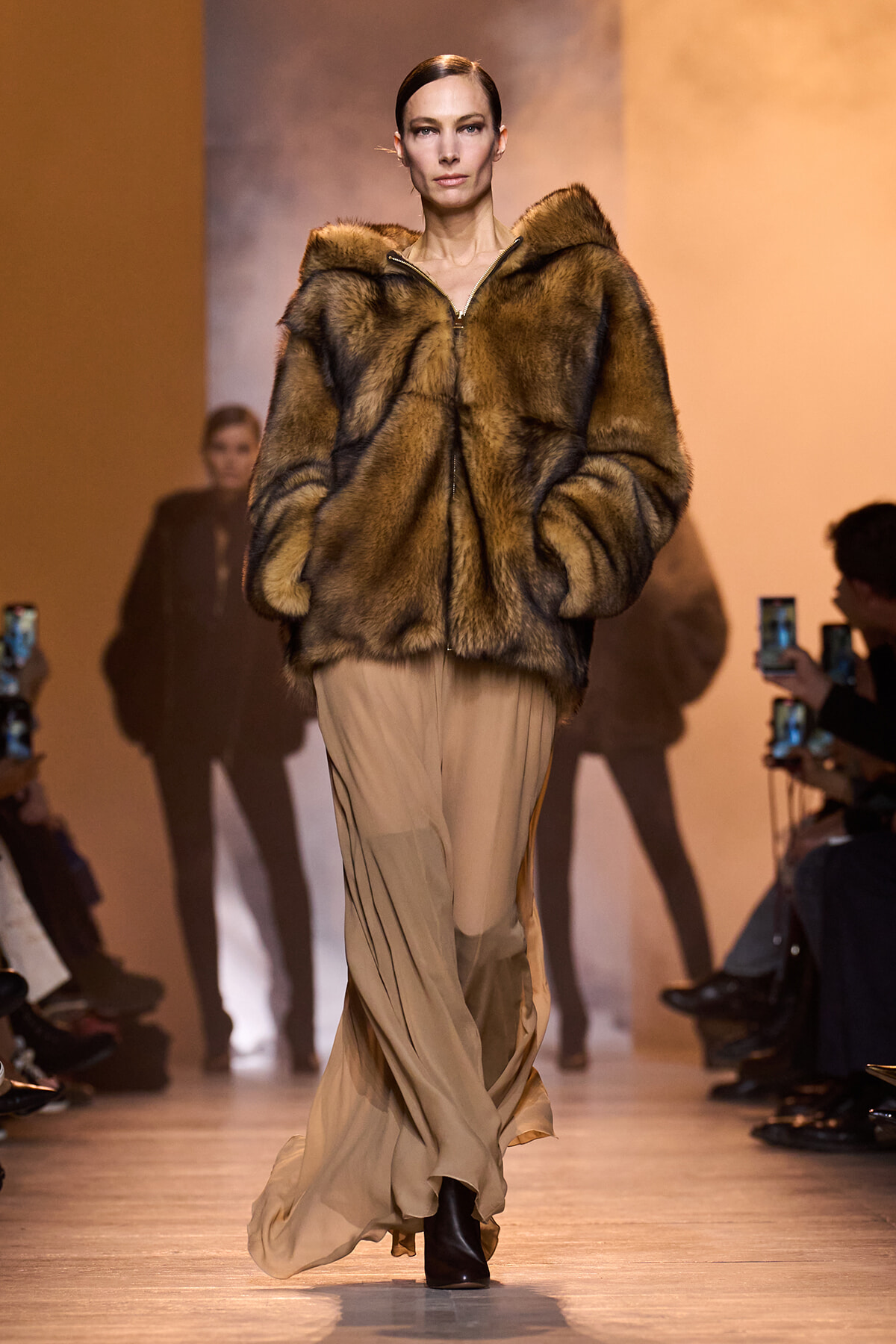 Model struts the runway wearing a brown fur coat over a beige, flowing dress with ankle boots; orange-lit backdrop behind her.