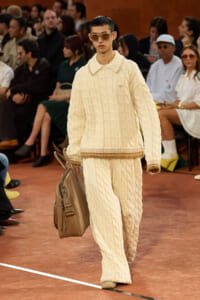 Model walks the runway in an oversized cream cable-knit sweater and matching knit pants, carrying a large tan leather bag.