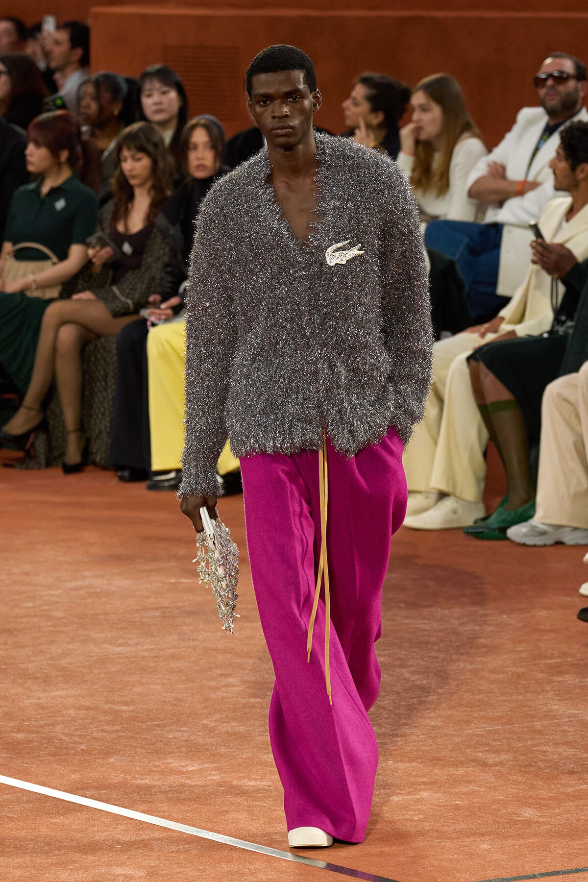 Male model on a runway wearing a sparkly silver fuzzy knit top and bright magenta wide-leg pants, carrying a beaded clutch among the seated audience.