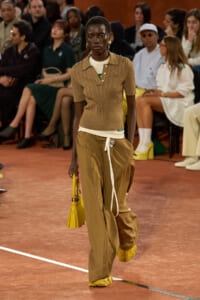Black female model walking a runway in a tan knit polo layered over a white shirt, oversized tan pants, yellow accessories, and a yellow handbag, with an audience watching.