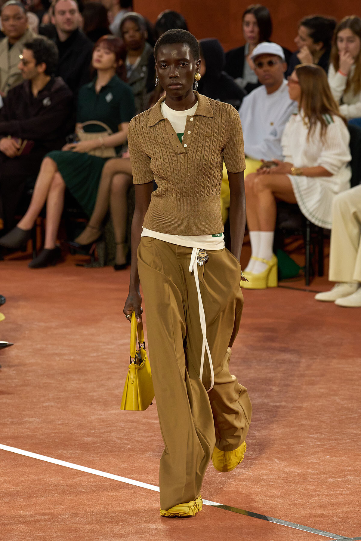 Black female model walking a runway in a tan knit polo layered over a white shirt, oversized tan pants, yellow accessories, and a yellow handbag, with an audience watching.