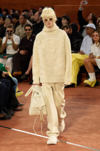 Model walking the runway in an all-cream knit turtleneck sweater and matching wide-leg pants, carrying a cream backpack and sunglasses, audience in background