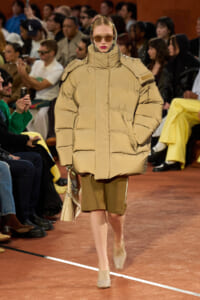 Model walks the runway wearing an oversized beige puffer coat with a tall collar, paired with beige footwear and a matching handbag, audience seated along the sides in the background.