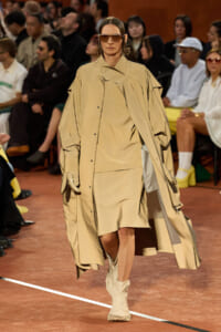 Model on a runway wearing an oversized beige trench coat with gloves and matching boots, sunglasses, as the audience watches in the background.