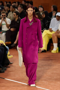 Model walking the runway in a bright magenta double-breasted coat with large front pockets and a decorative brooch.