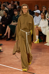 Model on a runway wearing a tan utility jumpsuit with large pockets, white belt, and yellow sneakers, carrying a tan bag.