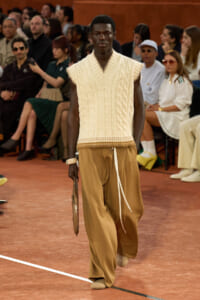 Male model on a fashion runway wearing a cream sleeveless cable-knit vest and camel wide-leg pants, holding a brown clutch as the audience watches.