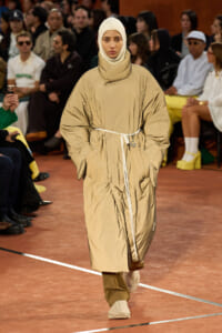 Model walks a runway wearing a tan oversized puffer coat with a belt, beige hijab, and matching sneakers.