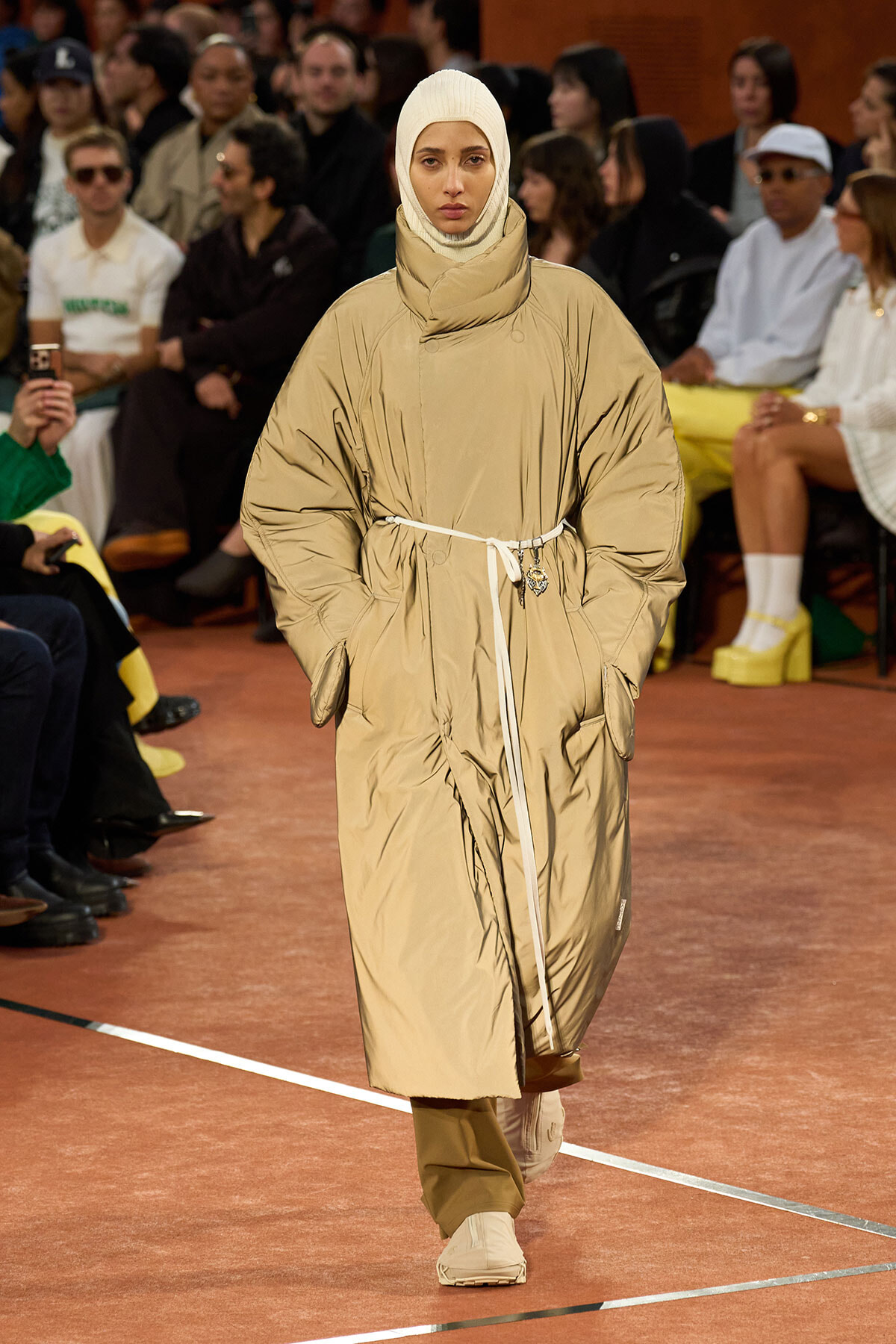Model walks a runway wearing a tan oversized puffer coat with a belt, beige hijab, and matching sneakers.