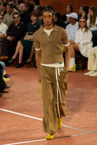 Male model on a runway wearing a brown knit polo tucked into tan wide-leg cargo pants, with yellow shoes and tinted sunglasses at a fashion show