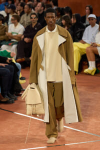 Male model strides down a runway in tan and cream layered coat, carrying a beige backpack as spectators sit along the catwalk.