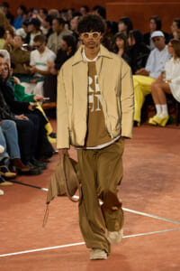 Runway model in a neutral-toned outfit: oversized tan pants, beige puffer jacket, white sunglasses, and a beige handbag walking the catwalk.