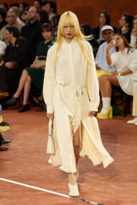 Model walks the runway in an ivory belted long-sleeve dress with a front slit; audience seated behind.
