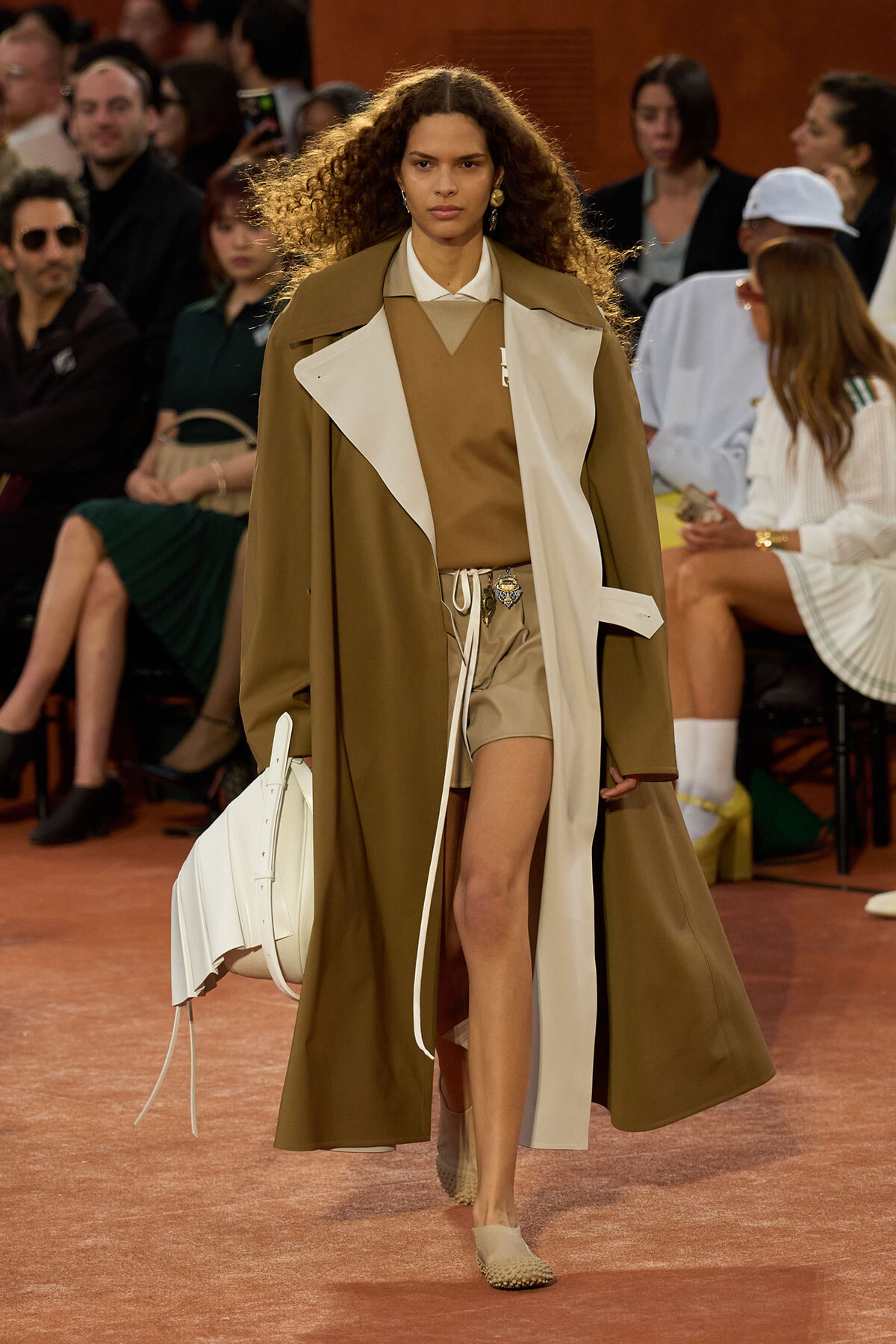 Model walks the runway in a camel oversized trench with cream lapels, paired with a beige top and shorts, carrying a large white handbag.