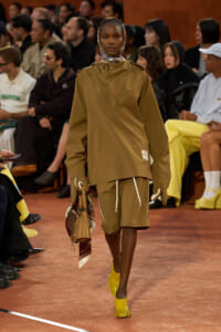 Model walking a fashion runway in a tan oversized top with matching shorts, yellow heels, and a headscarf, carrying a tan handbag.