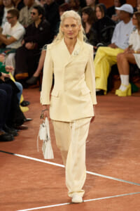 Model walking the runway in a cream double-breasted suit with a matching bag as the audience watches.